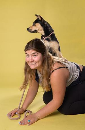 A woman with long brown hair is kneeling on a yellow background, smiling at the camera. A small black and tan dog is sitting on her back, looking to the side.