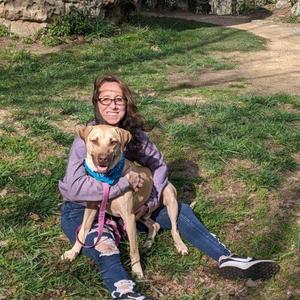 A woman with glasses is sitting on the grass with a dog on her lap. The dog is wearing a blue bandana and a pink leash. They are both looking at the camera.